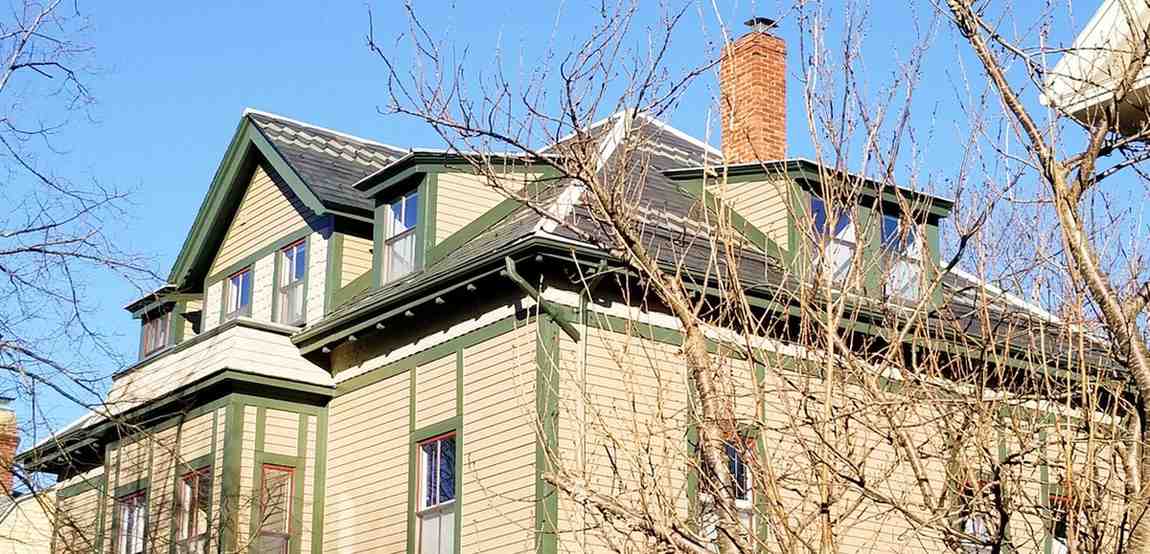 Victorian-style house with green trim and tan siding viewed through bare winter branches. A red brick chimney contrasts against the clear blue sky.