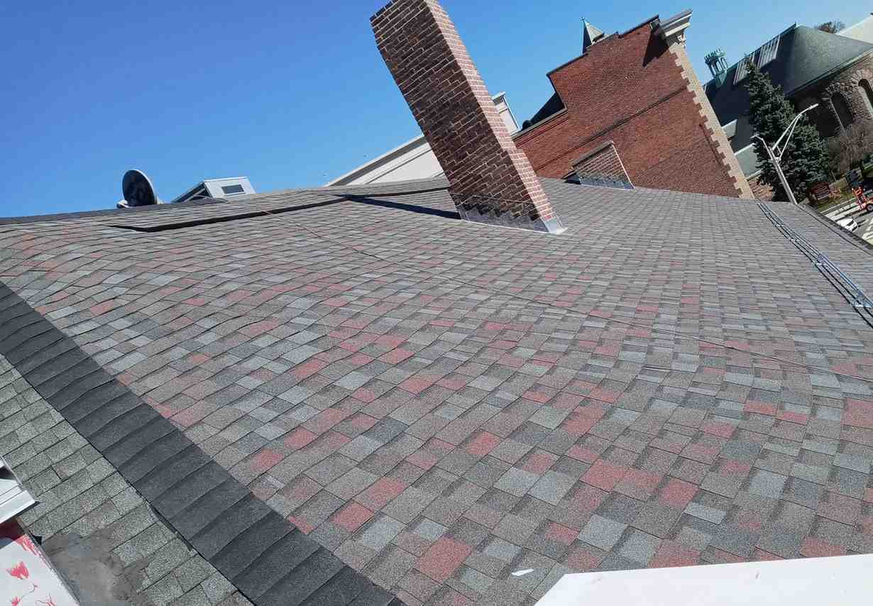 A rooftop with red and gray shingles under a clear blue sky, featuring a brick chimney. Nearby, a brick building and green trees are visible.